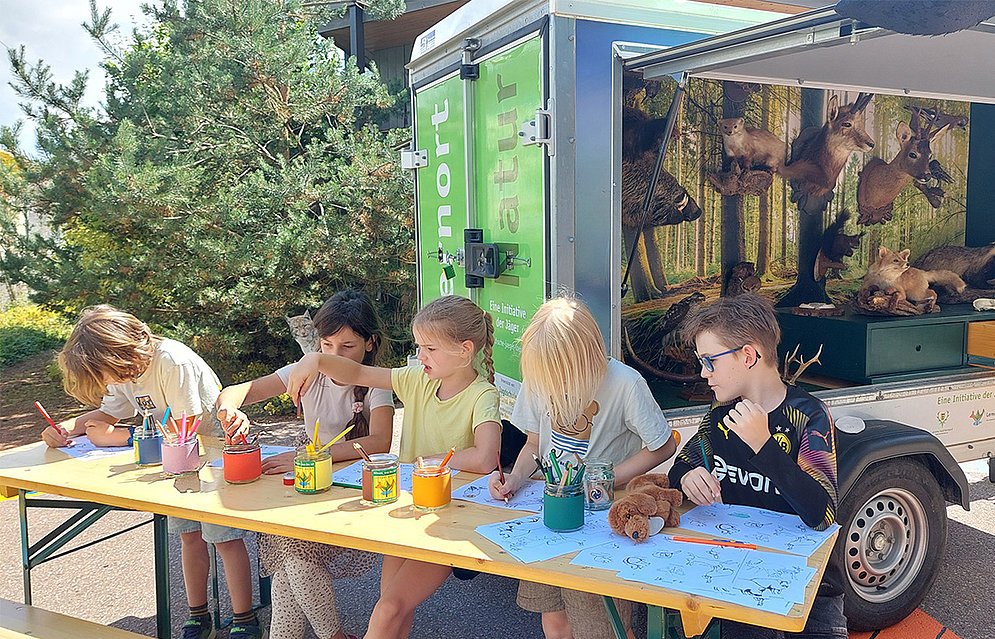 Jäger Max Vogel zu Besuch an der Schöpflin Schule mit seinem Info-Mobil »Lernort Natur«, Kinder sitzen an einem Tisch und zeichnen. Im Hintergrund ist das Info-Mobil mit Tierfellen zu sehen.  