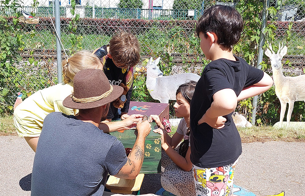 Jäger Max Vogel zu Besuch an der Schöpflin Schule mit seinem Info-Mobil »Lernort Natur«. Kinder sitzen um einen Tisch herum und hören Max Vogel zu.  